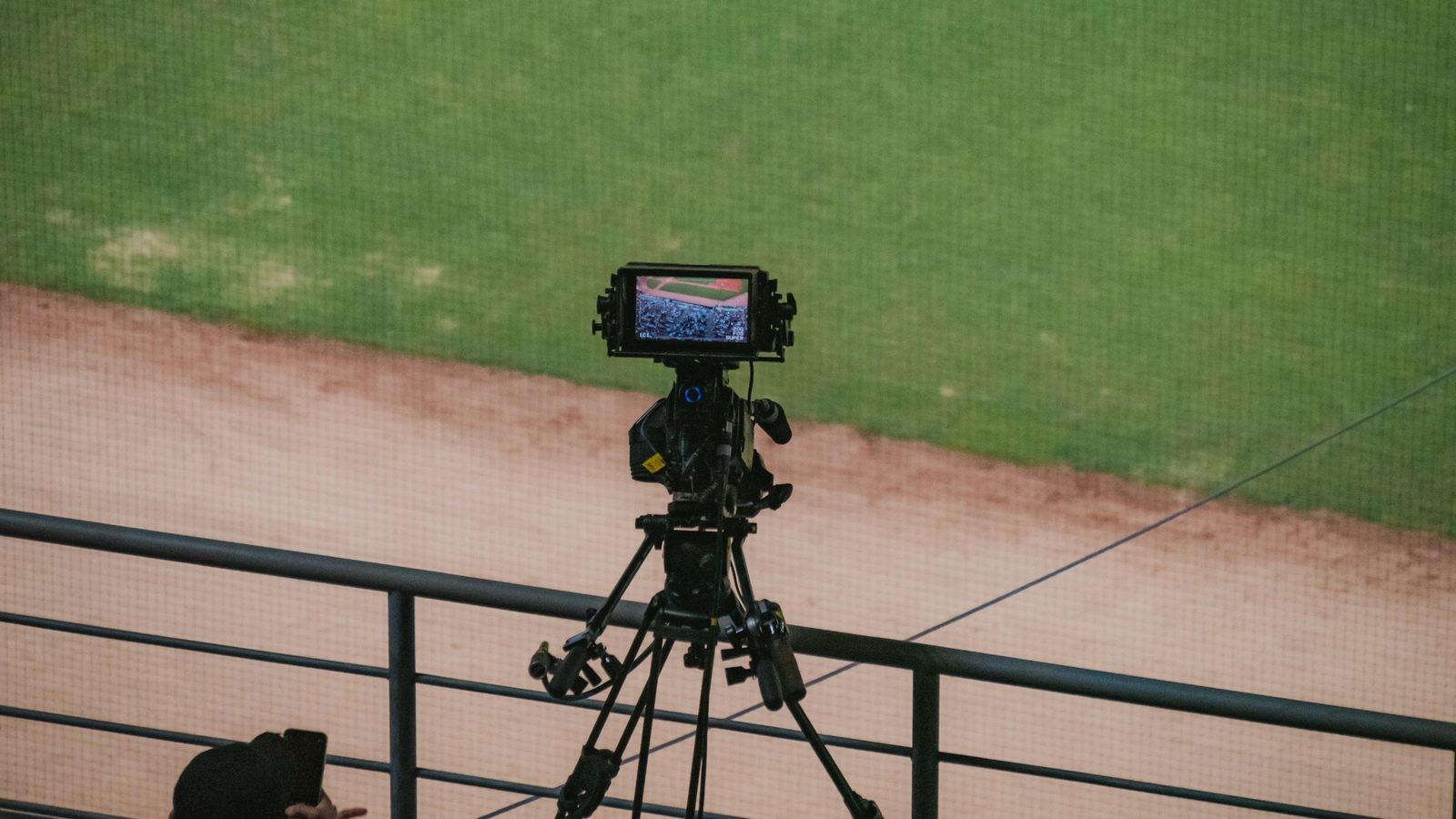A camera on a tripod on the rail above a baseball field, pointed at the infield