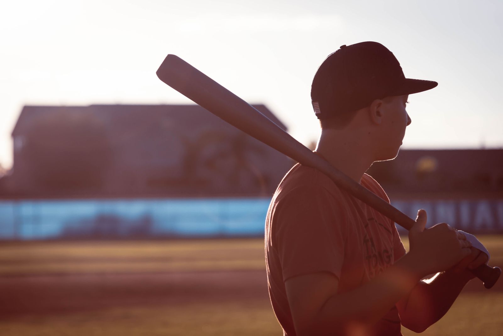 Teenage player resting a bat on his shoulder at a field in late-afternoon light
